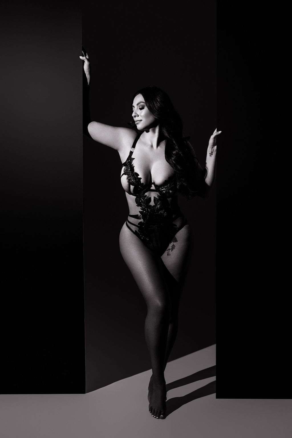 A woman poses barefoot in a black lace bodysuit against a dark background, with one arm raised and the other resting on a panel. The photo is in black and white.