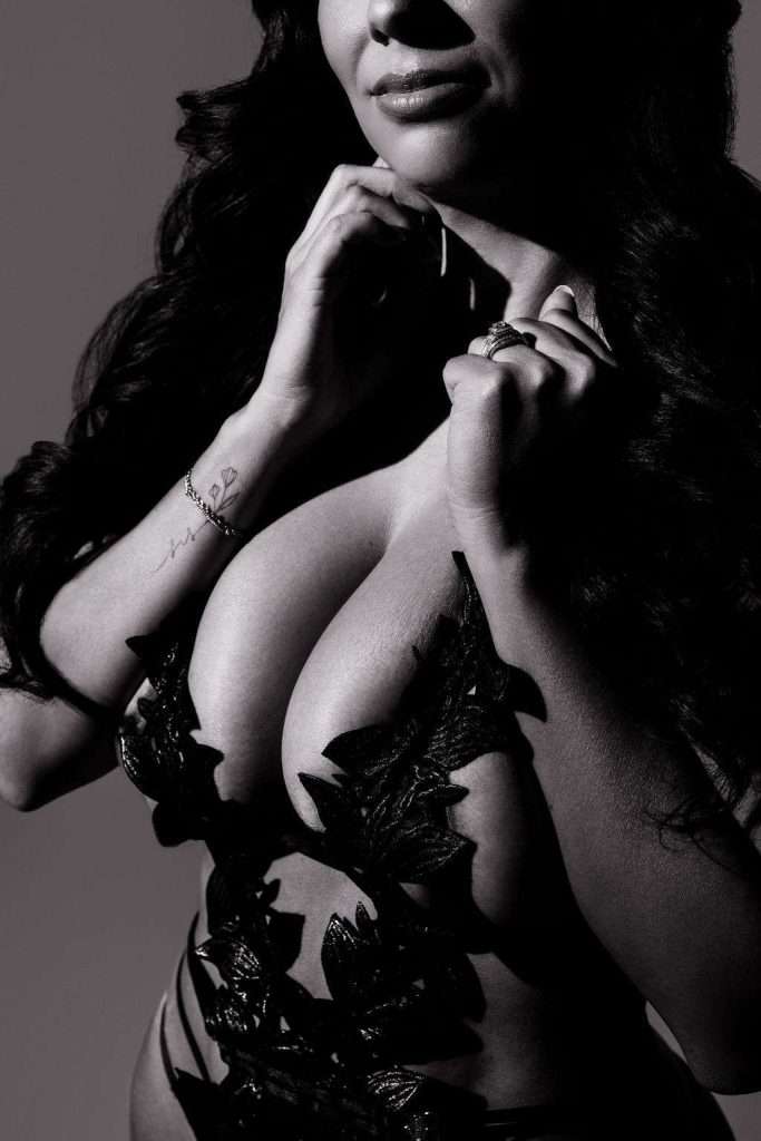 A woman in black lingerie with leaf patterns poses in dramatic lighting, her hands near her neck and face partially obscured by shadow.