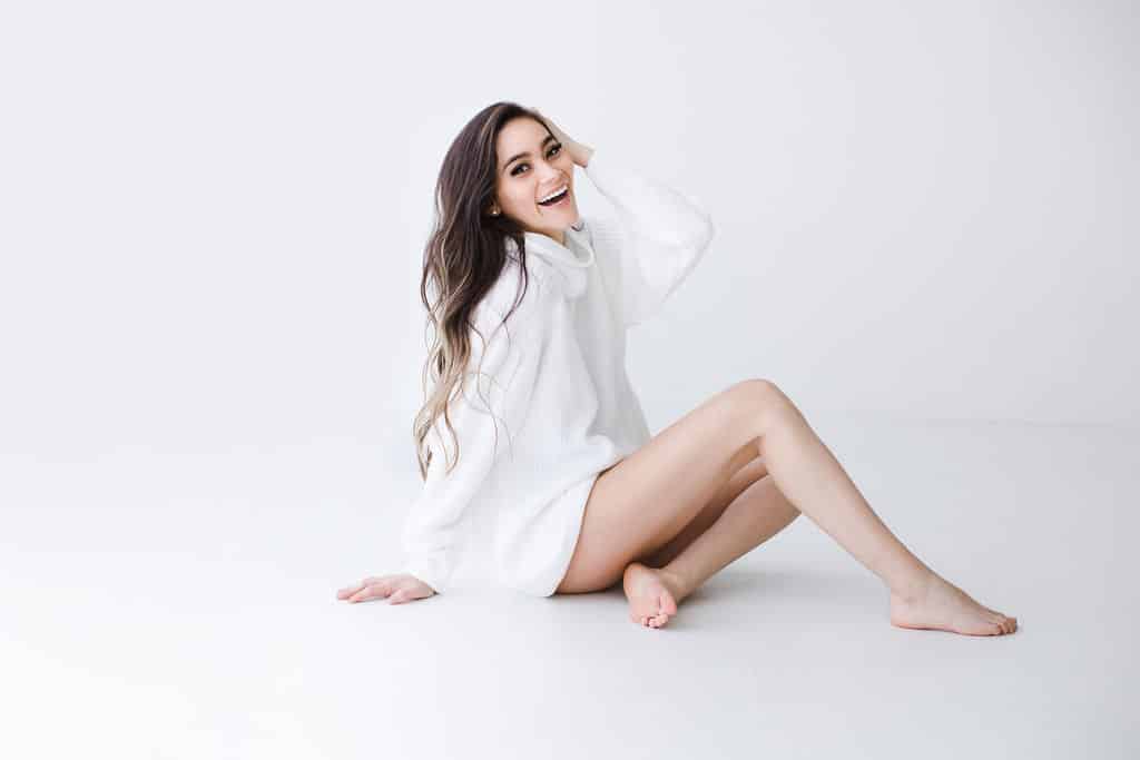 Woman with long brown hair sits on the floor against a white background, wearing an oversized white sweater and smiling at the camera.