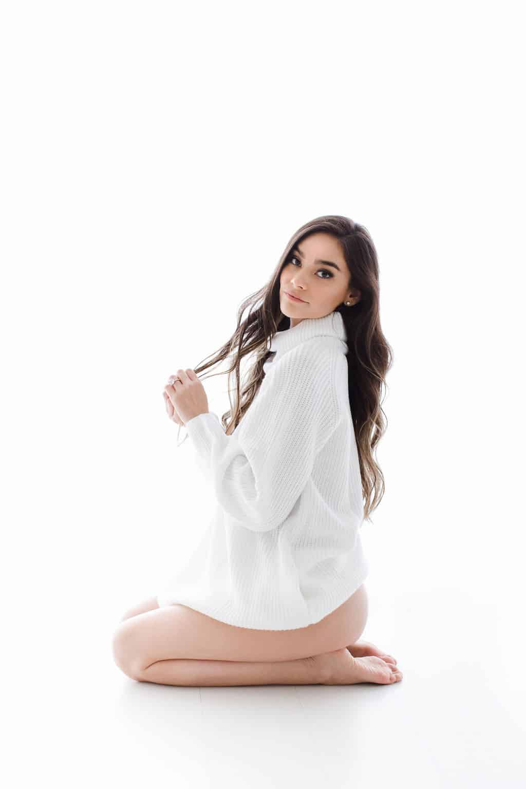 A woman with long brown hair kneels on the floor, wearing an oversized white sweater against a white background.
