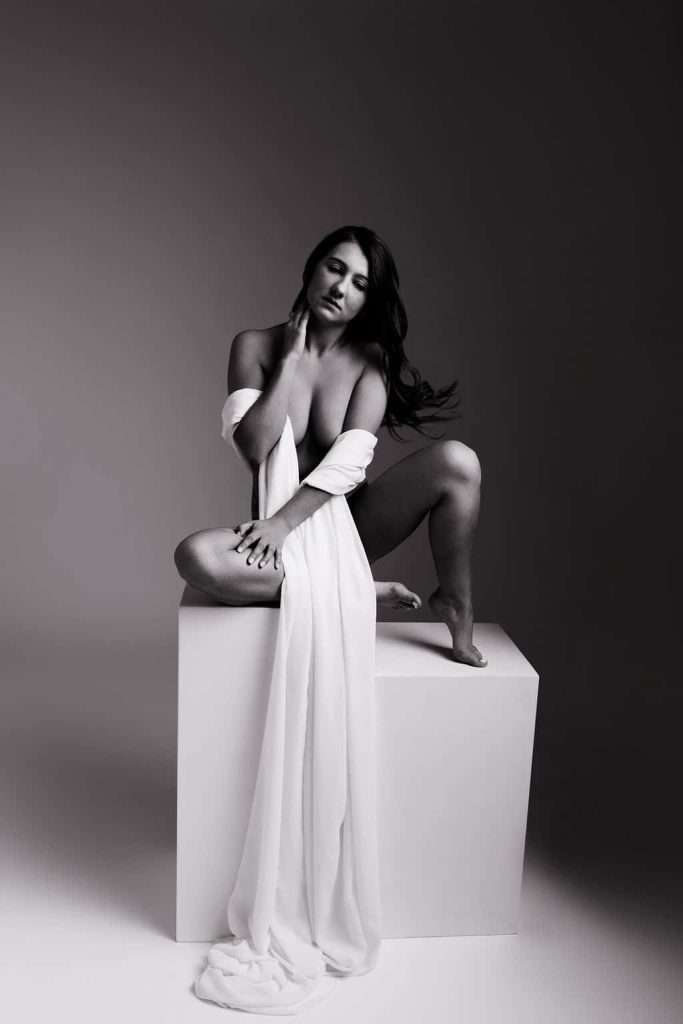 A woman draped in a long white fabric sits on a cube, with one knee raised and hand touching her neck, in a black and white studio setting.