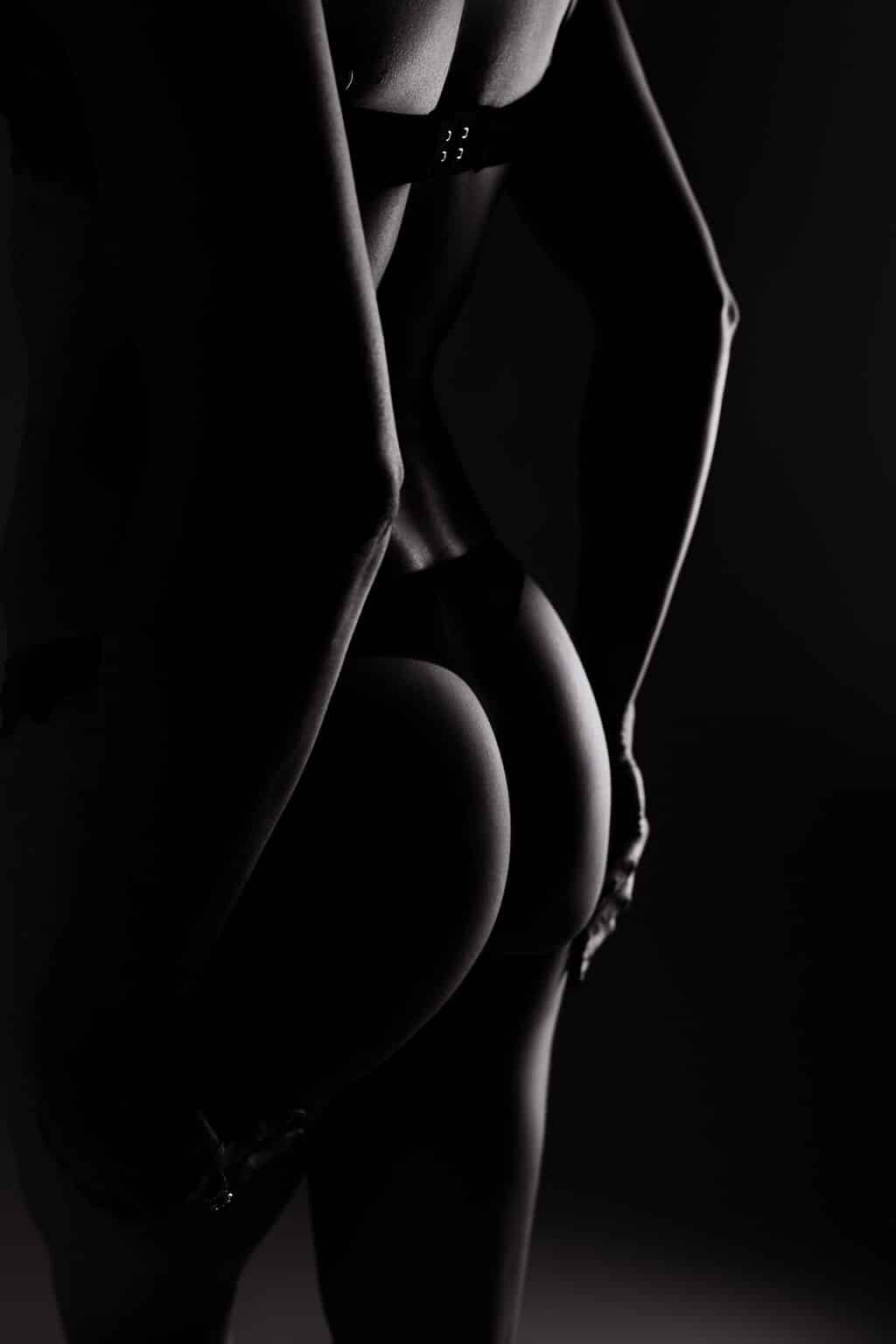 A black and white photo showing the back view of a person wearing lingerie, with emphasis on their lower back and buttocks in dramatic lighting.