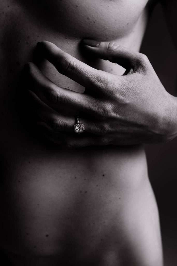 A close-up, black and white photo of a hand with an engagement ring resting on a bare torso.