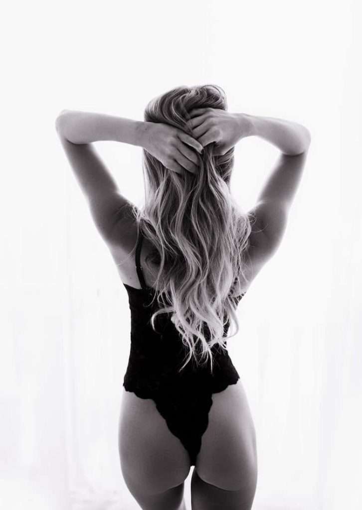 A woman with long hair stands with her back to the camera, wearing a black lace bodysuit and holding her hair up with both hands.