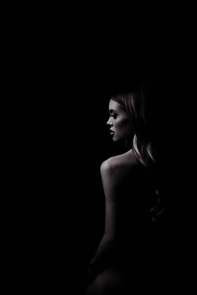 Black and white photo of a woman with long hair, shown from the back and side in low lighting, facing to the right against a dark background.