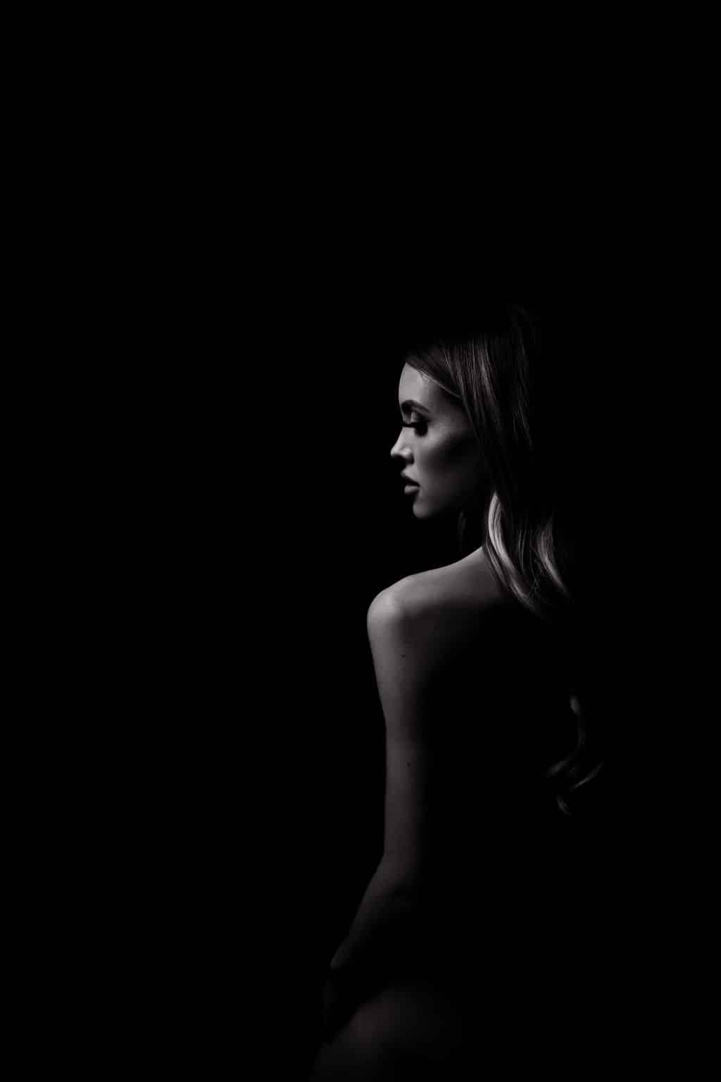 Black and white photo of a woman with long hair, shown from the back and side in low lighting, facing to the right against a dark background.