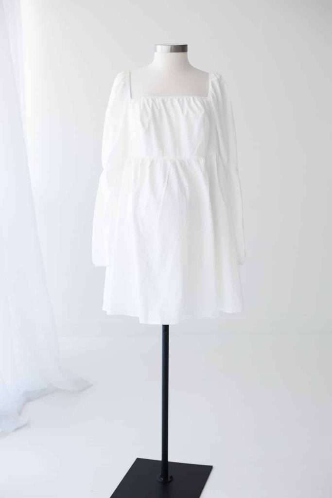 A white long-sleeve, knee-length dress with a square neckline is displayed on a dress form against a plain white background.
