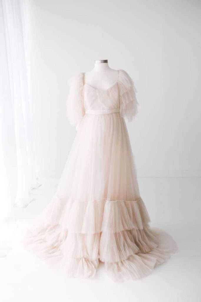 A light pink, floor-length tulle gown with ruffled tiers and puffed short sleeves displayed on a dress form in a white room.