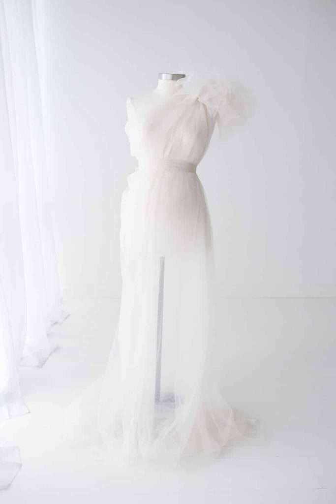 A white mannequin displays a sheer, floor-length, one-shoulder tulle dress in a minimalistic, white-walled room with soft natural light.