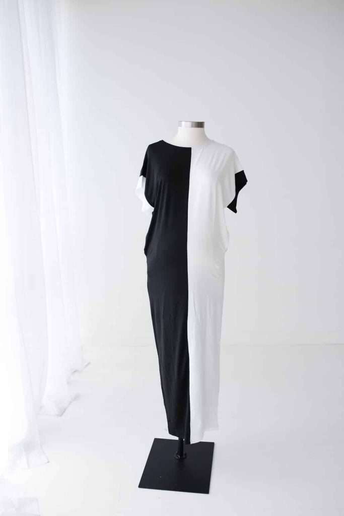 A mannequin displays a sleeveless, ankle-length dress with a vertical black and white color split in a bright, minimalist room.