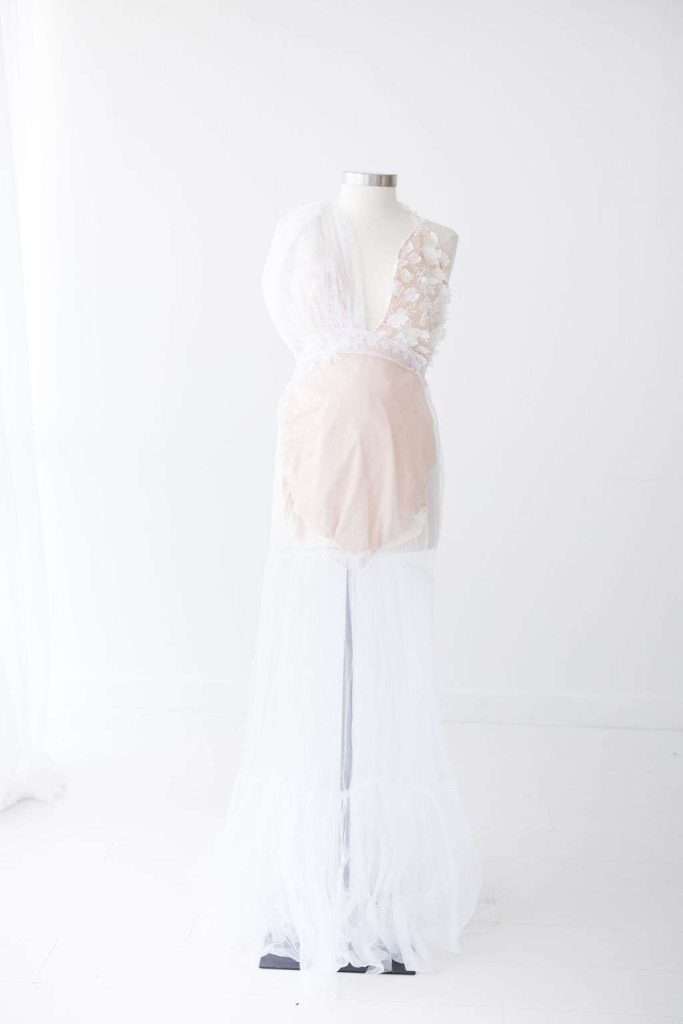 A white mannequin displays a sheer white gown with lace and floral details on the bodice, set against a plain white background.