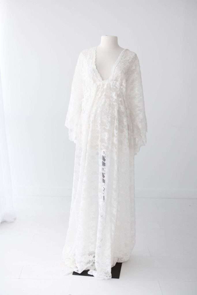 A long, white, sheer lace dress with a deep V-neck and loose sleeves displayed on a headless dress form in a white studio.