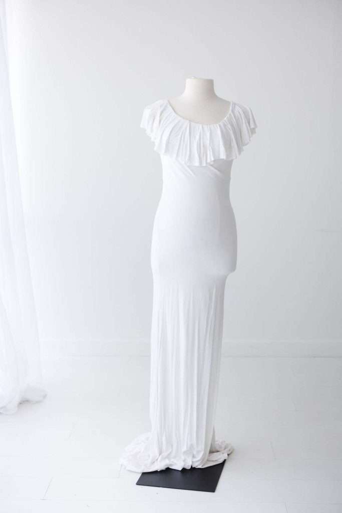 A white, floor-length dress with short sleeves and a ruffled neckline is displayed on a mannequin in a minimal, white room.