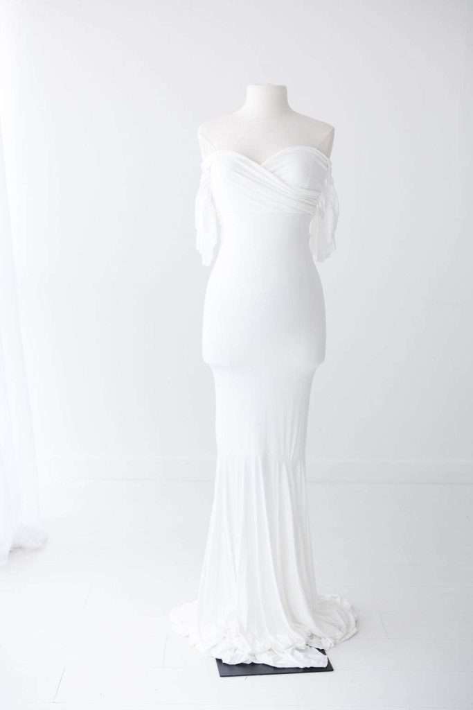 A floor-length, off-the-shoulder white gown is displayed on a dress form in a minimal, white-walled room.