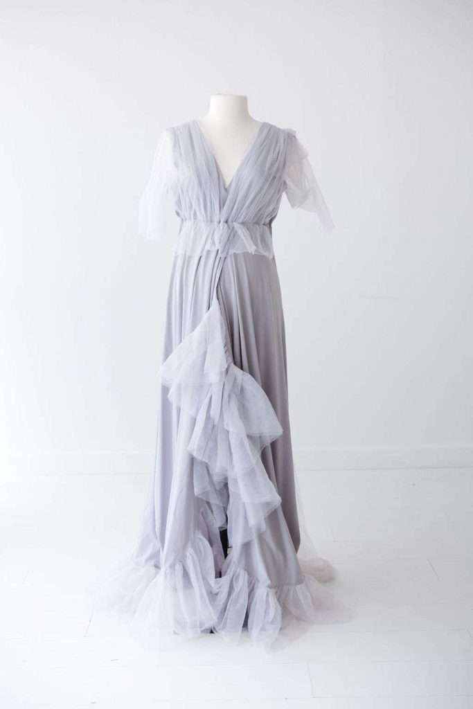 Light gray floor-length gown with a deep V-neck, sheer flutter sleeves, and layers of ruffled tulle cascading down the front, displayed on a white dress form against a plain background.