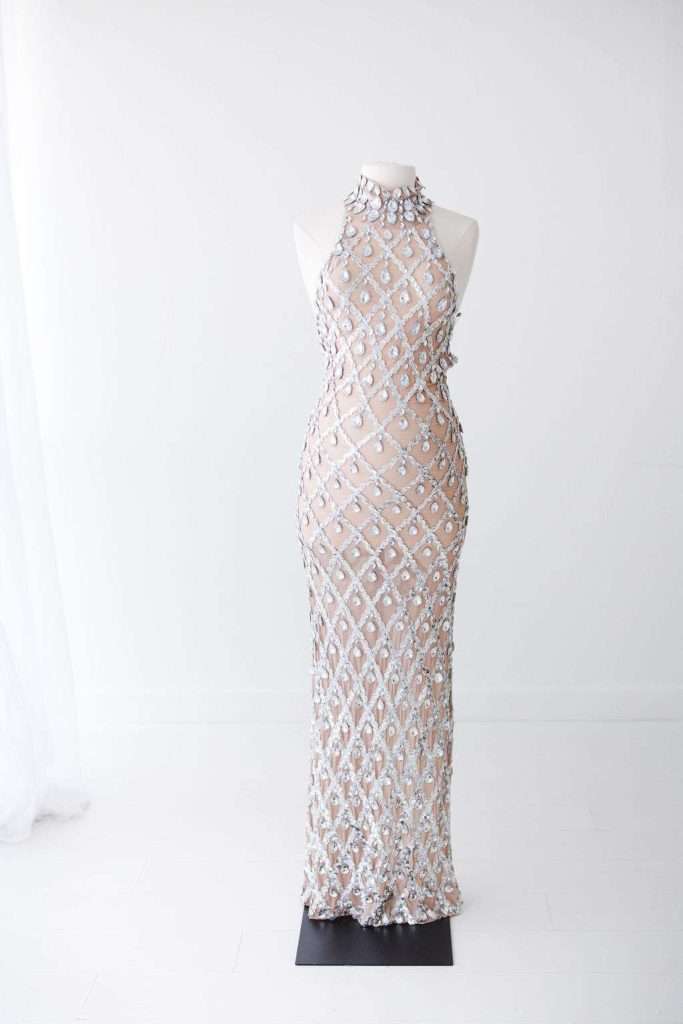 A floor-length, sleeveless nude gown with a high neckline, covered in a silver geometric beaded pattern, displayed on a dress form against a white background.