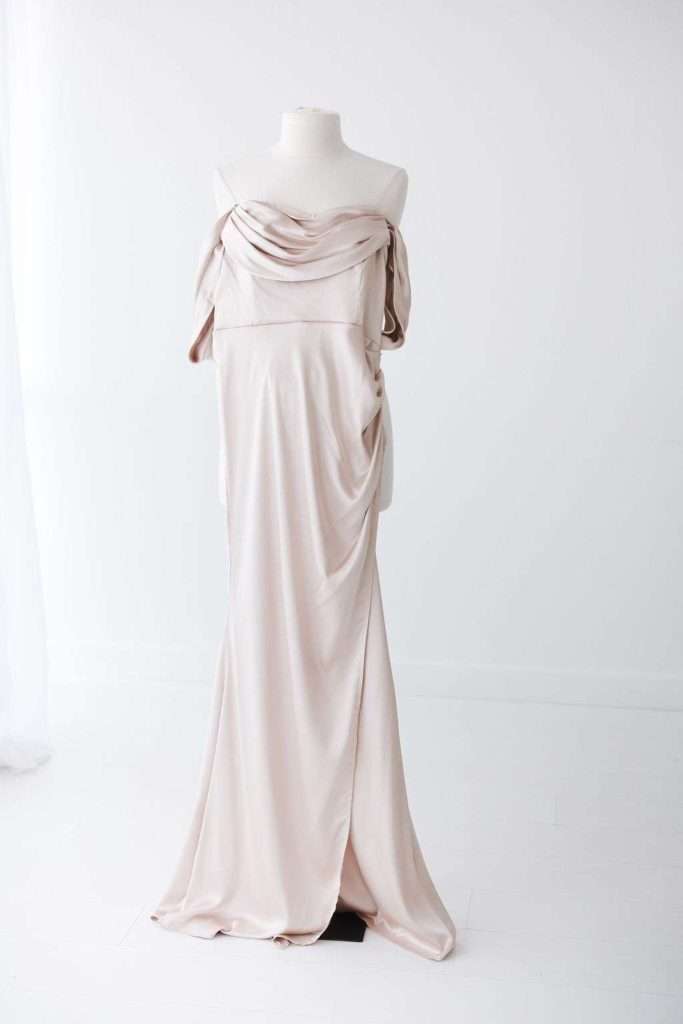 A cream-colored, off-the-shoulder satin gown displayed on a dress form in a minimalist, white room.