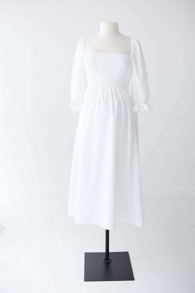 A white midi dress with puffed long sleeves, smocked bodice, and square neckline displayed on a mannequin against a plain white background.