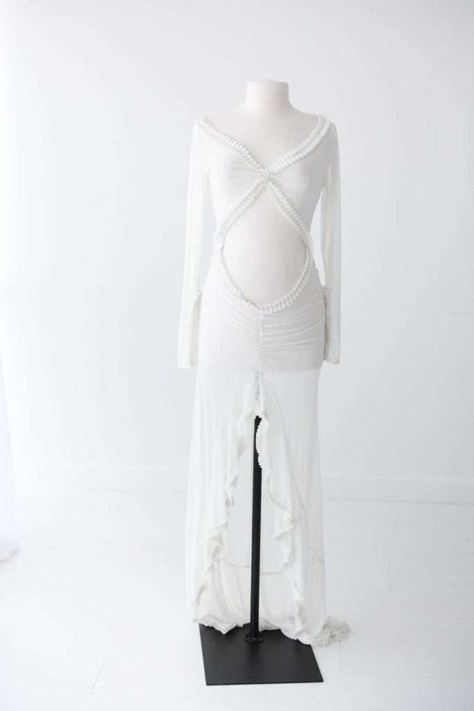 White mannequin displays a long-sleeved, white maternity gown with pearl trim, crisscross bodice, open belly design, and ruffled hem detail, set in a plain white room.