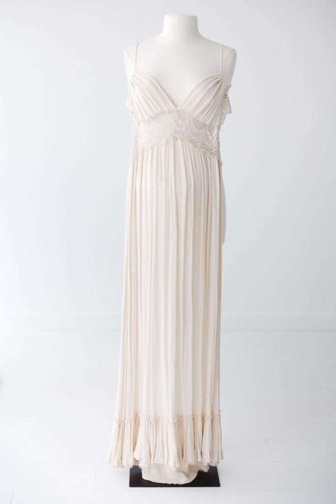 A cream-colored, floor-length sleeveless dress with a V-neckline, decorative beading at the bodice, and a ruffled hem, displayed on a mannequin against a plain background.