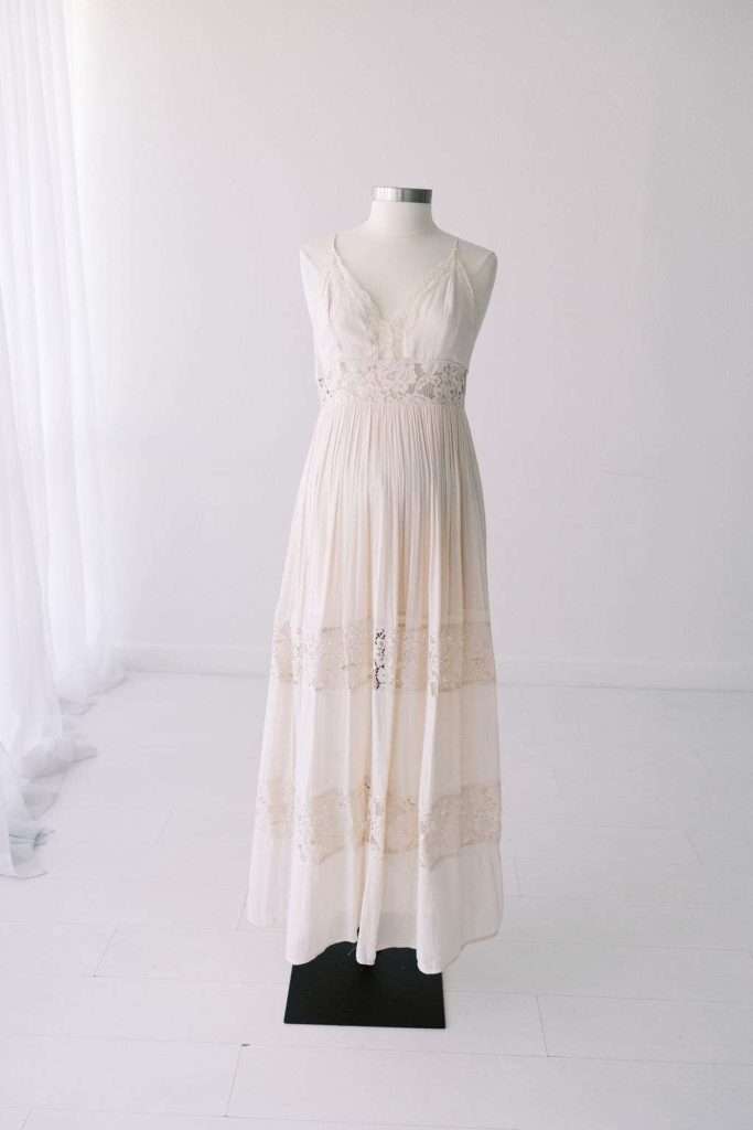 A white sleeveless, floor-length dress with lace details is displayed on a mannequin in a minimal, light-filled room.
