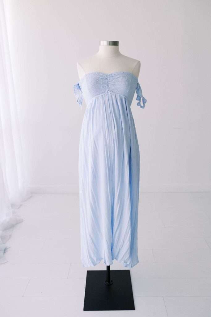 Light blue off-shoulder maxi dress displayed on a dress form in a minimal, white-walled room with sheer curtains.