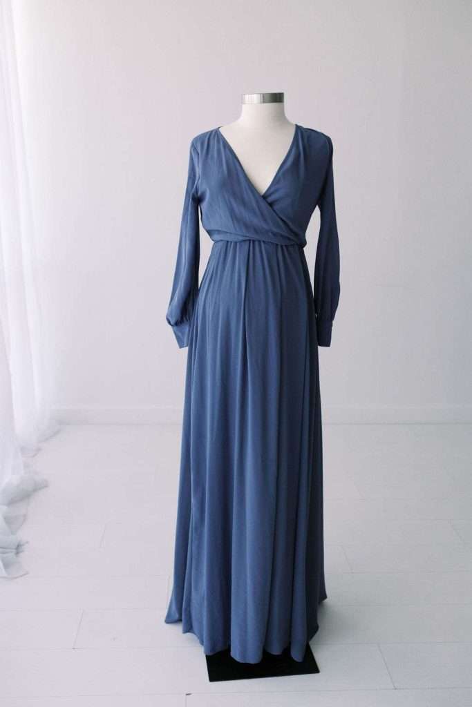 A mannequin displays a long, blue, V-neck dress with long sleeves and a gathered waist, set against a plain white background.