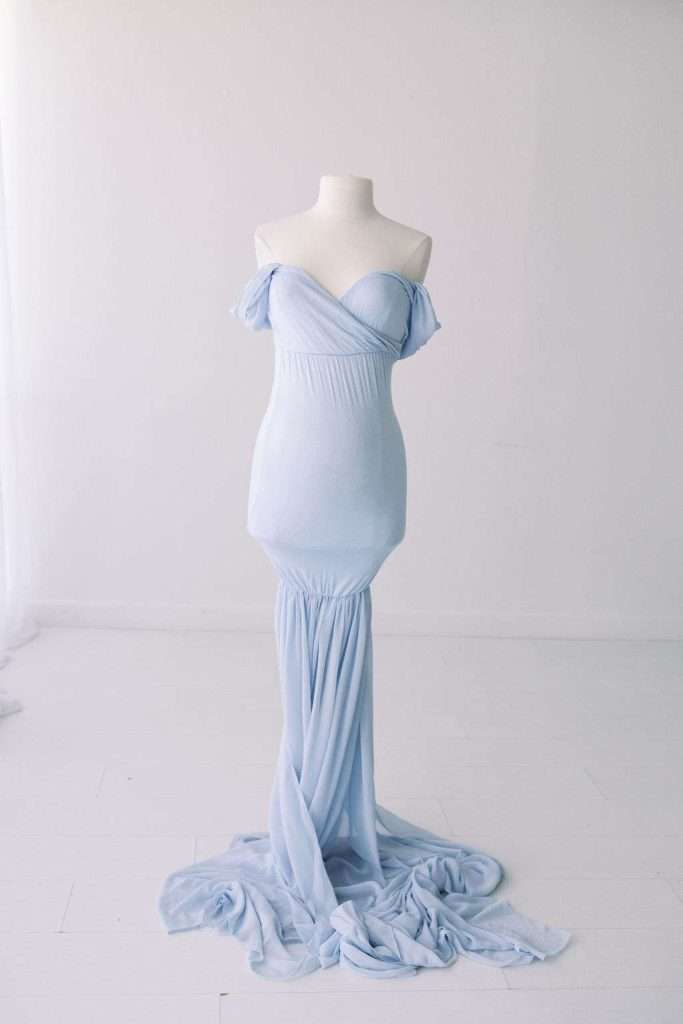 A light blue off-shoulder gown is displayed on a white dress form against a plain white background, with fabric pooling on the floor.