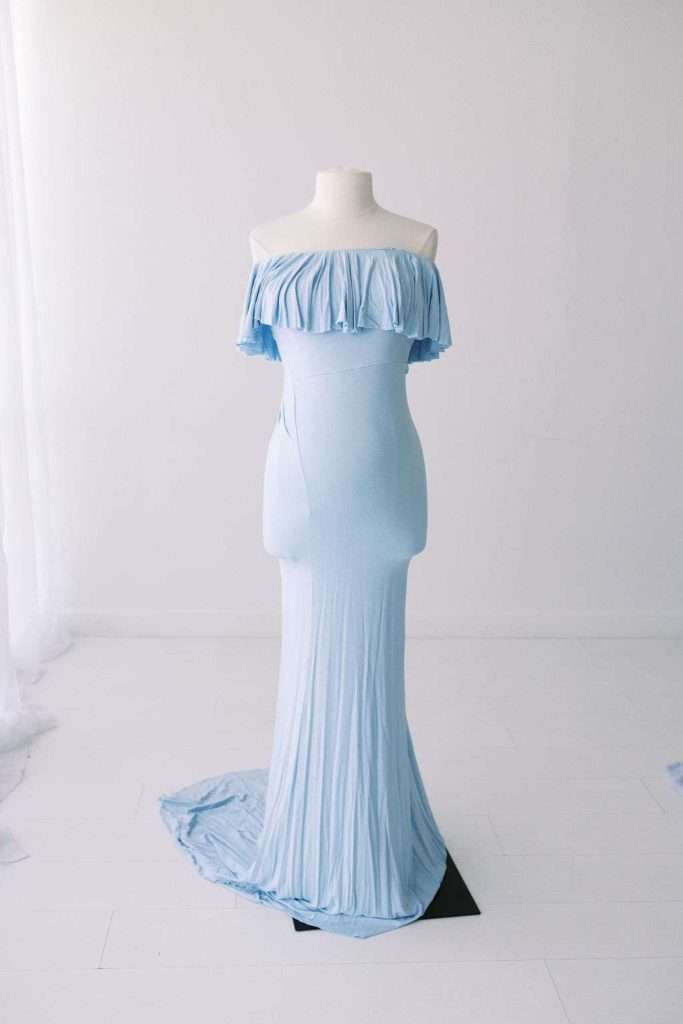 A light blue, off-the-shoulder, floor-length dress displayed on a white dress form in a minimal studio setting.