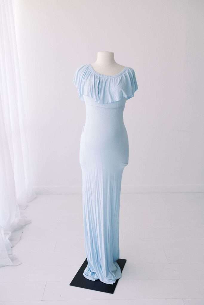 Light blue, floor-length dress with ruffled off-shoulder neckline displayed on a white dress form in a minimal, white room.