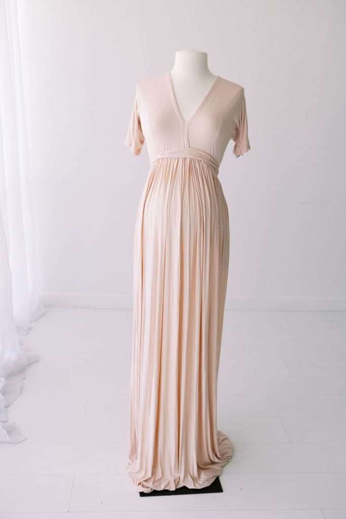 A floor-length, short-sleeve, light blush maternity dress is displayed on a mannequin in a white room.