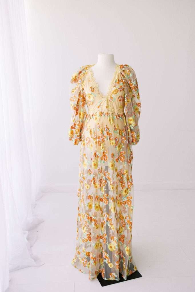 A mannequin displays a long, sheer floral dress with puffed sleeves and a deep V-neck, set in a minimal white room with soft natural light.
