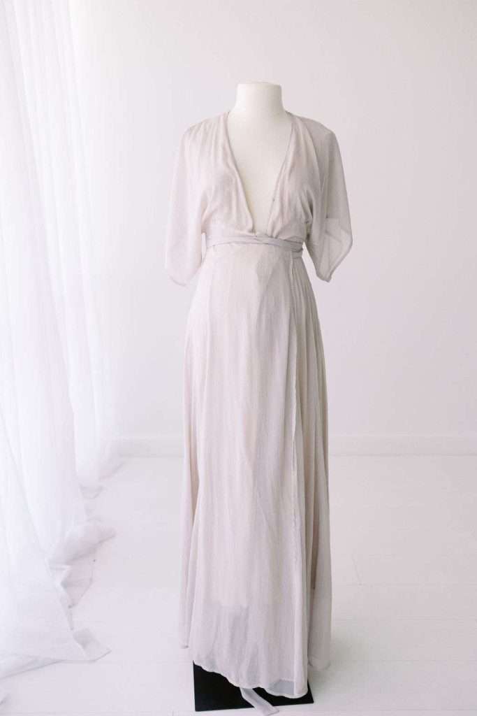 A white, floor-length dress with short sleeves and a deep V-neck displayed on a mannequin in a bright, minimalist room.