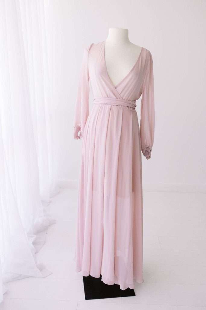 Light pink, long-sleeve, floor-length dress with a deep V neckline and a fitted waist, displayed on a white mannequin in a minimal, bright room.