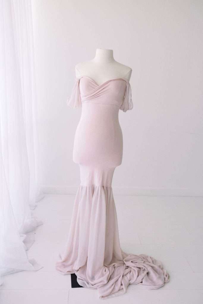 A strapless, light pink mermaid-style gown is displayed on a mannequin in a white, minimalist room with sheer curtains.