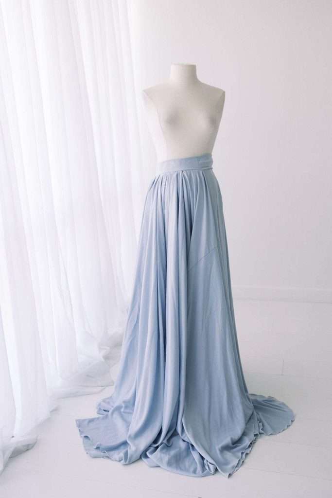 A white mannequin displays a long, flowing, light blue skirt in a bright, minimalist room with sheer white curtains.