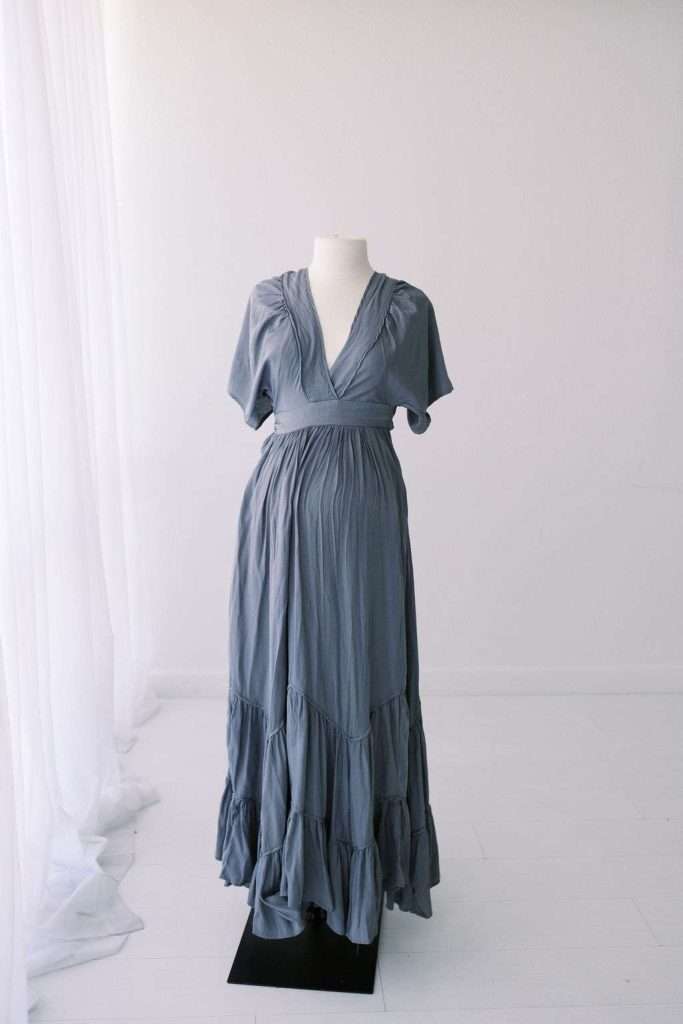 A gray, short-sleeved, V-neck maxi dress with a gathered waist and tiered skirt is displayed on a headless mannequin in a bright, minimal room.