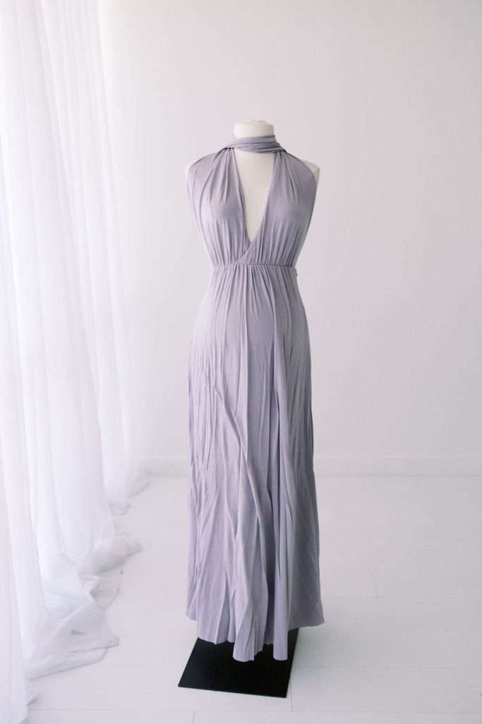 A sleeveless, floor-length lavender dress with a halter neckline displayed on a dress form in a white room with sheer curtains.