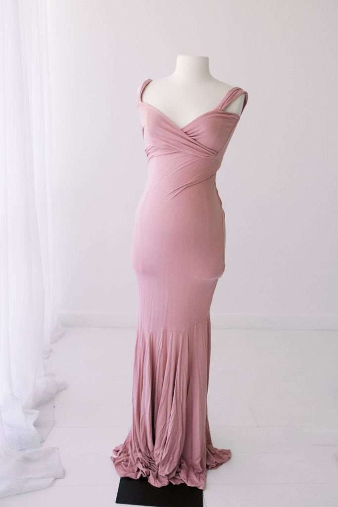 Mannequin displaying a floor-length, fitted, dusty pink gown with gathered straps and a ruched, draped bottom in a bright, minimal studio setting.