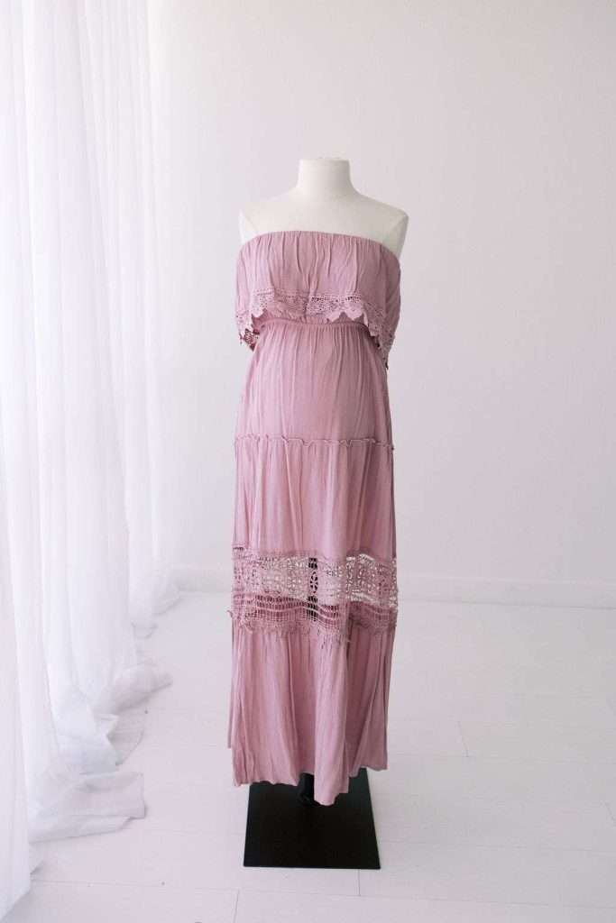 A mauve off-shoulder maxi dress with lace detailing is displayed on a mannequin in a white, minimalist room with sheer curtains.