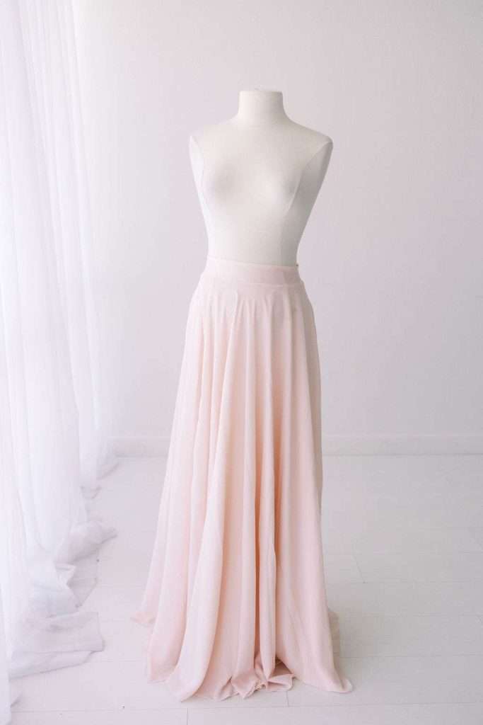 A white mannequin displays a long, flowing, pale pink skirt in a minimalist, softly lit room with sheer white curtains.