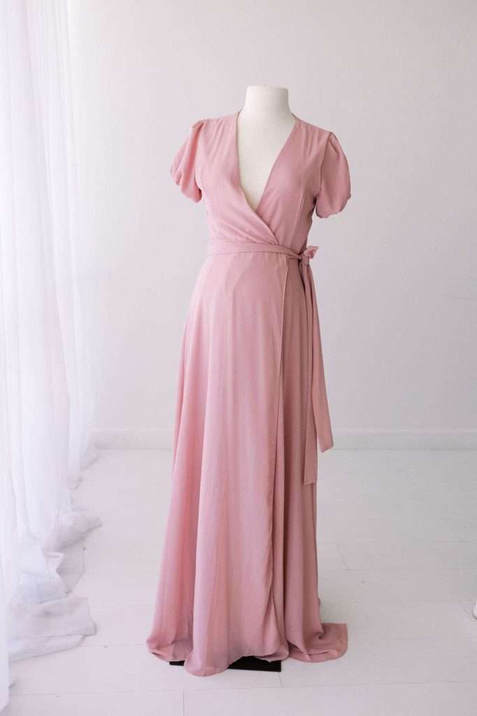 A long, light pink wrap dress with short puff sleeves and a V-neck is displayed on a white mannequin in a bright, minimal room.