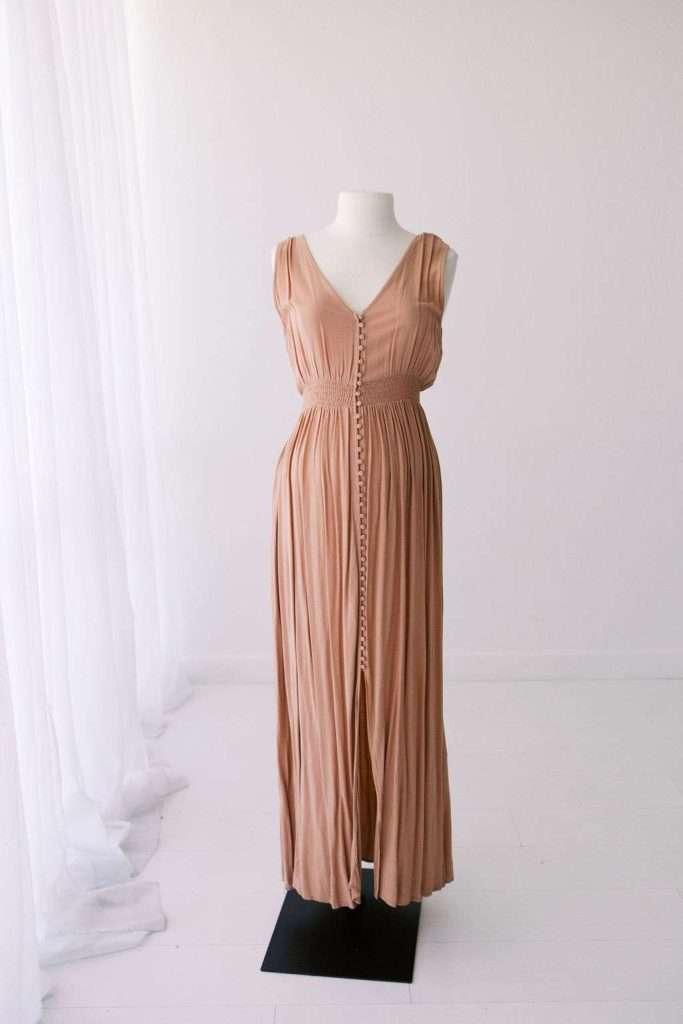 Sleeveless, light brown pleated maxi dress with front buttons displayed on a white mannequin in a minimal studio with white walls and curtains.