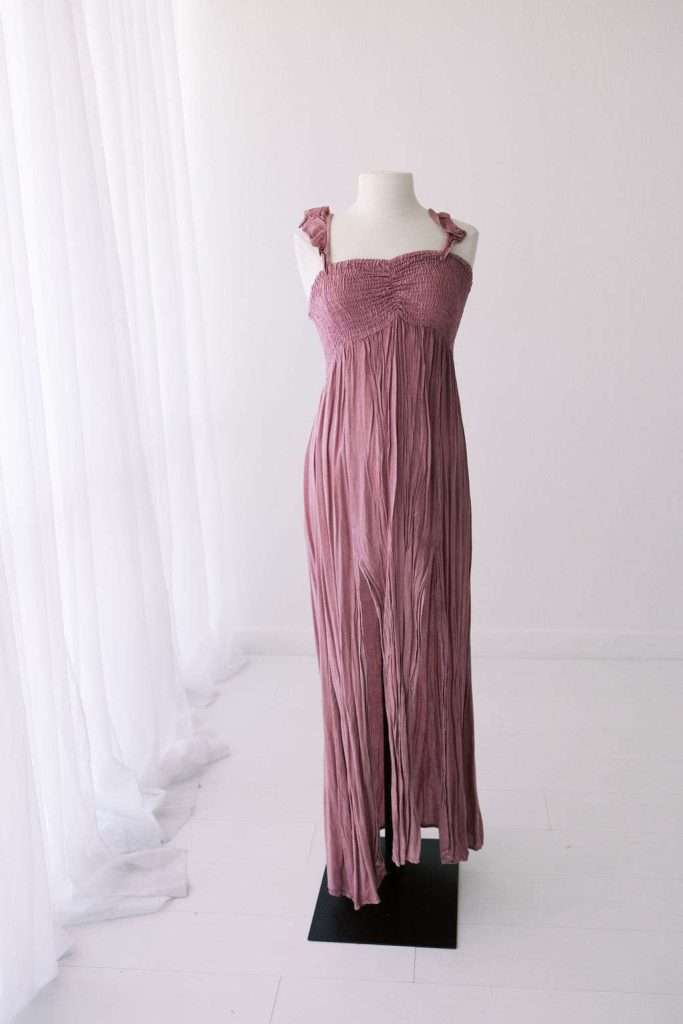 A mauve, sleeveless maxi dress with knotted shoulder straps and a smocked bodice is displayed on a mannequin in a minimal, white room with sheer curtains.