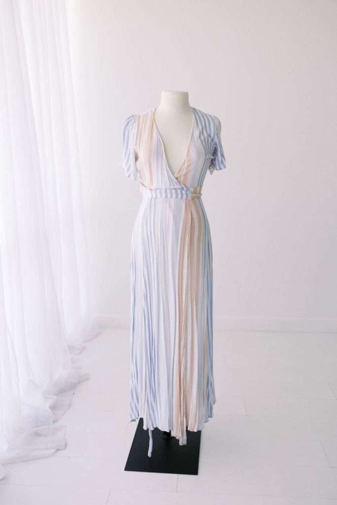 A white mannequin displays a light, short-sleeve, V-neck, ankle-length dress with vertical pastel stripes, set in a minimal, white room with sheer curtains.