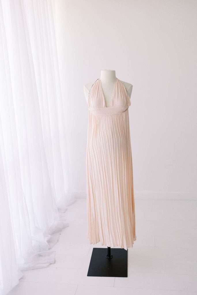Sleeveless, light pink pleated halter dress displayed on a mannequin in a bright, minimal room with sheer white curtains.