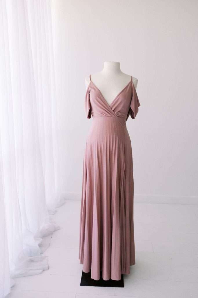 A mauve, floor-length dress with off-the-shoulder straps displayed on a white mannequin in a minimalist, white room with sheer curtains.