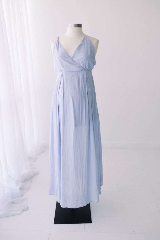 A light blue sleeveless, floor-length dress with a V-neckline is displayed on a mannequin in a minimal, white room.