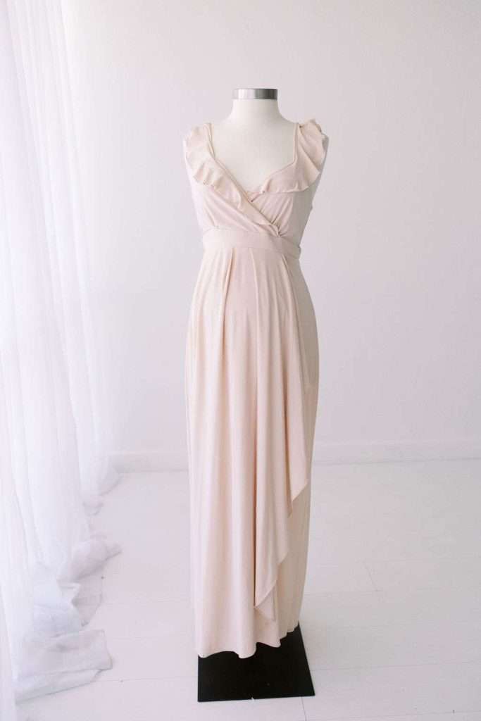 Sleeveless, light pink floor-length dress with ruffled straps displayed on a dress form in a bright, minimal room with white floors and sheer curtains.