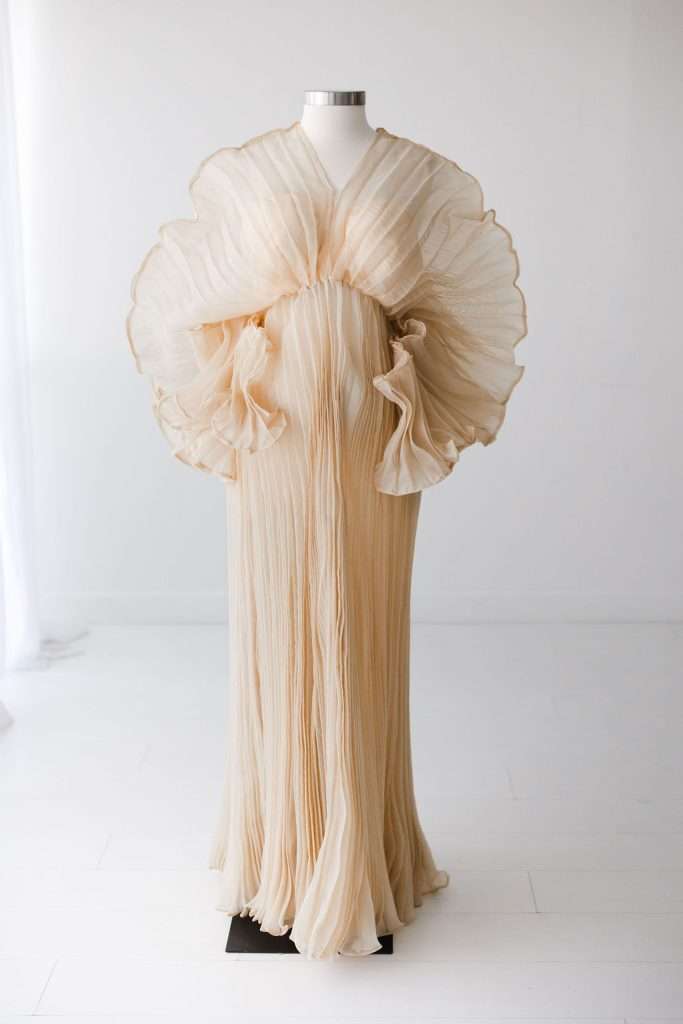 Ivory pleated gown with large, ruffled organza details around the shoulders and chest, displayed on a dress form in a minimalist white room.